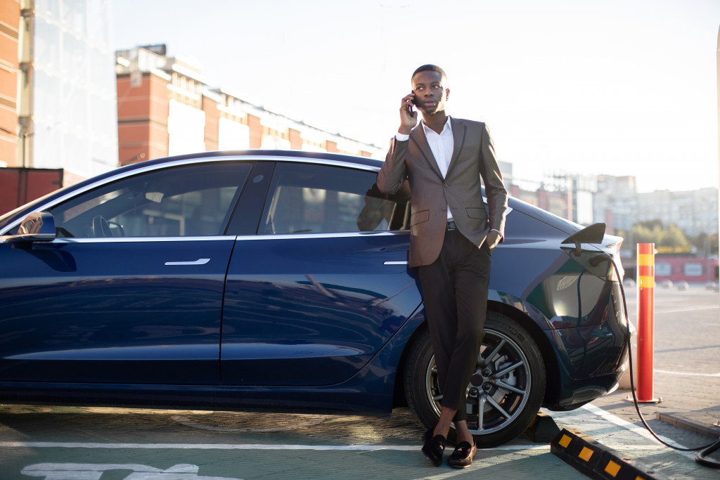 A man in a suit talks on the phone while leaning on his electric car at a charging station